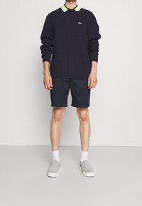 Jersey de punto de cable en color navy con un cuello en amarillo pálido y verde, combinado con pantalones cortos navy y zapatillas grises sin cordones. Diseño limpio y sencillo.