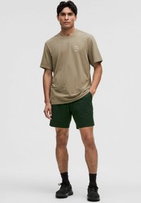 Beige bomullst-shirt med kort ärm, i kombination med mörkgröna shorts. Modellen står självsäkert med händerna i fickorna och har på sig svarta sneakers.