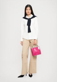 Donna che indossa una camicia bianca con un maglione blu scuro sulle spalle, pantaloni beige a gamba larga, tacchi neri e tiene in mano una borsa fucsia.