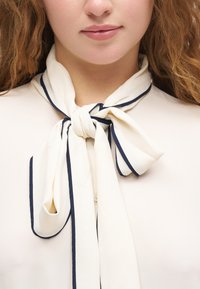 Žena nosi krémovú blúzku s veľkým golierom s mašľou lemovaným tmavým lemom, záber zblízka na výstrih a pery.