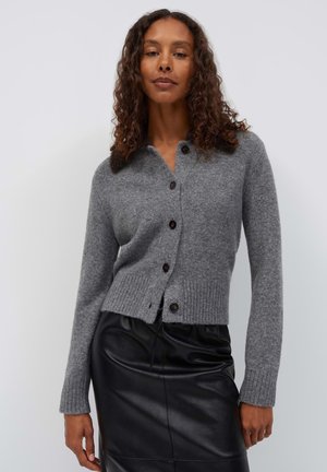 Donna con capelli ricci che indossa un cardigan grigio abbottonato e una gonna nera in pelle, in piedi davanti a uno sfondo chiaro e semplice.
