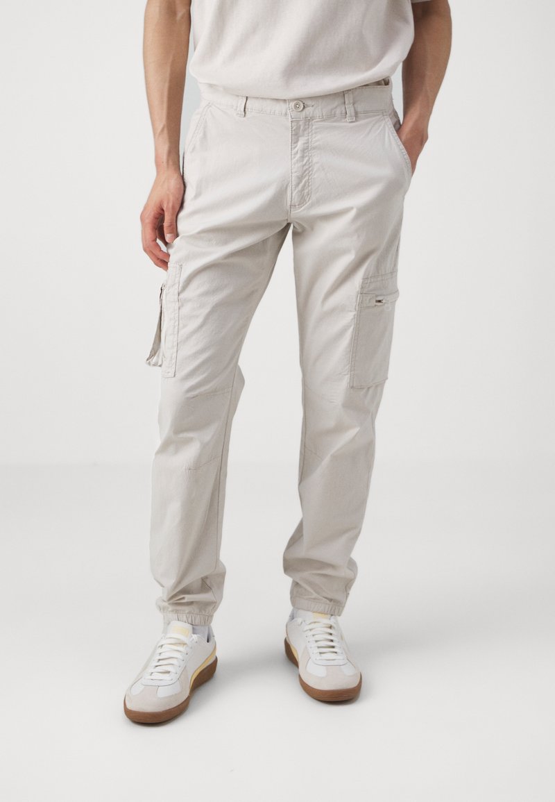 Mężczyzna w jasnobeżowych spodniach cargo z kieszeniami i białych trampkach z gumową podeszwą, stojący na tle białego tła.