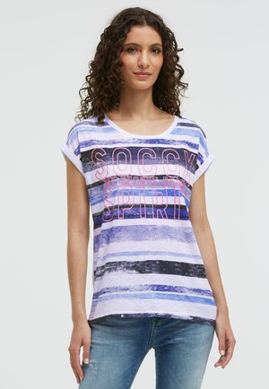 Frau mit lockigem Haar, die ein blau-weiß gestreiftes T-Shirt mit pinkem Text und blaue Jeans trägt, steht vor einem schlichten hellen Hintergrund.