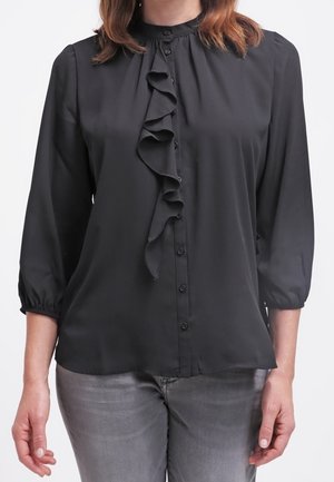 Mujer con blusa negra de manga larga con detalle de volantes en la parte delantera y vaqueros grises, de pie frente a un fondo blanco.