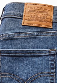 Vaqueros de mezclilla con un lavado azul medio, que presentan una parche de cuero marrón con el logo, costuras visibles y un clásico diseño de cinco bolsillos.