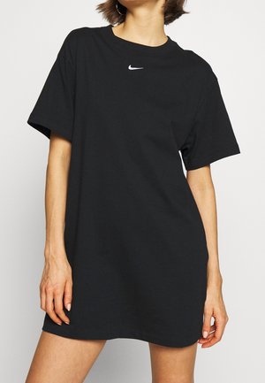 Femme portant une robe T-shirt noire Nike ample à manches courtes avec un petit logo blanc sur la poitrine, debout devant un fond uni.