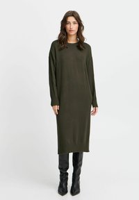 Abito lungo in maglia verde oliva con polsini e orlo a coste, abbinato a stivali neri aderenti fino al ginocchio. Tessuto morbido, vestibilità rilassata.