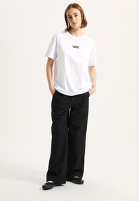 White, short-sleeve t-shirt with black Vans logo, paired with loose-fitting black trousers and flat black shoes. Plain background.