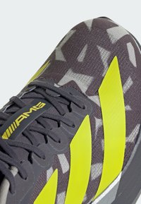 Nahaufnahme eines dunkelgrauen Sneakers mit schwarzen Schnürsenkeln, neon-gelben Streifen und AMG-Logo auf der Zunge, mit geometrisch gemustertem Stoff.