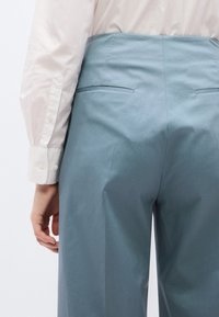 Lichtblauwe broek van gladde stof; heeft een hoge tailleband en twee achterzakken, gecombineerd met een wit overhemd met lange mouwen en knopen.