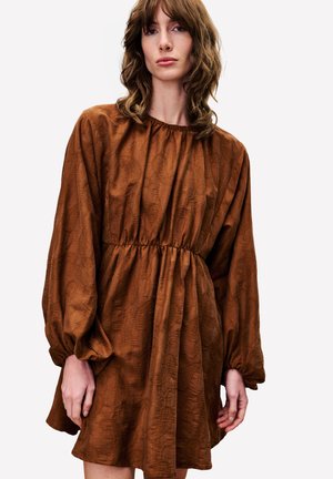 Frau mit welligem braunem Haar, die ein rostfarbenes, langärmeliges Kleid mit gerafftem Bund und subtilem Texturmuster auf einem weißen Hintergrund trägt.