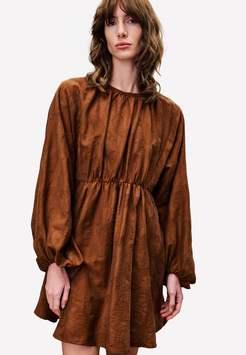 Frau mit welligem braunem Haar, die ein rostfarbenes, langärmeliges Kleid mit gerafftem Bund und subtilem Texturmuster auf einem weißen Hintergrund trägt.