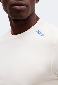 Κοντινό πλάνο ενός ατόμου που φοράει λευκό πουκάμισο με μπλε κείμενο "BORG" στον αριστερό ώμο, εμφανίζονται μόνο ο λαιμός και το κάτω μέρος του προσώπου.