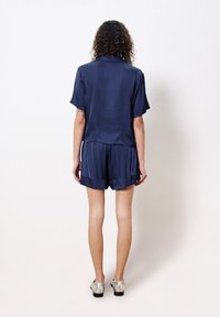 Marineblaue Satin-Shortsleeve-Hemd und passendes Shorts-Set mit lockerer Passform, das eine glatte Textur und einen dezenten Glanz aufweist. Silberne Schuhe runden den Look ab.