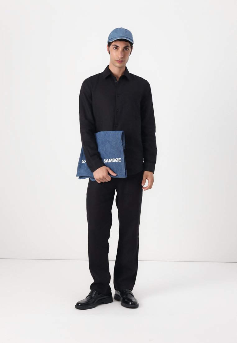 Chemise noire à boutons, pantalon noir et chaussures noires. Tient un carnet en denim bleu avec "SAMSOE" en texte blanc. Casquette bleue. Fond blanc.