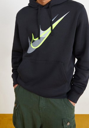 Person trägt einen schwarzen Kapuzenpullover mit überlappenden grauen, weißen und neongelben Nike-Swoosh-Logos, kombiniert mit grünen Cargohosen.
