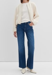 En krämfärgad, fluffig bomberjacka över en vit topp, kombinerad med högmidjade blåjeans. Modellen bär spetsiga vita klackar. En enkel, avslappnad outfit.