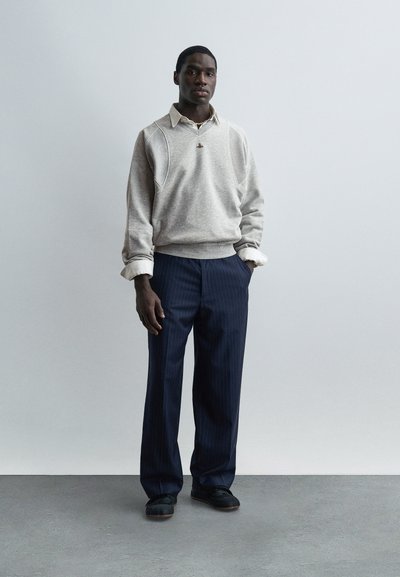 Άνδρας που φοράει ανοιχτό γκρι πουλόβερ πάνω από πουκάμισο με γιακά, μπλε σκούρο παντελόνι με λεπτές ρίγες και μαύρα παπούτσια, στέκεται με τα χέρια στις τσέπες μπροστά σε απλό φόντο.