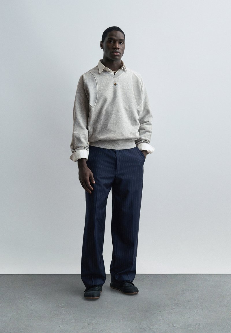 Uomo con maglione grigio chiaro sopra una camicia con colletto, pantaloni a righe blu navy e scarpe nere, in piedi con le mani nelle tasche contro uno sfondo semplice.