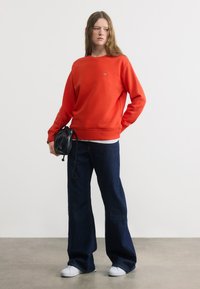 Γυναίκα με κόκκινο φούτερ, σκούρο παντελόνι φαρδιάς γραμμής, λευκά αθλητικά παπούτσια, κρατώντας μια μικρή μαύρη τσάντα, στέκεται μπροστά σε απλό φωτεινό φόντο.