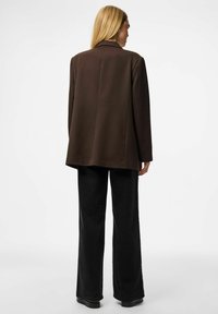 Rjav oversized blazer s sodobnim krojem, z reverjem in brez vidnih zaponk, v kombinaciji s širokimi črnimi hlačami. Gladka tekstura.