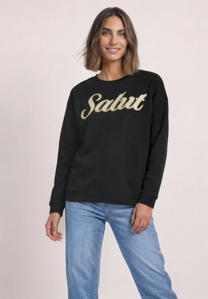 Femme aux cheveux bruns mi-longs souriante portant un sweat-shirt noir avec le texte "Salut" et un jean bleu clair sur un fond uni.
