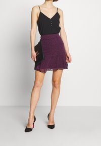 Zwart mouwloos topje met knoopdetails, gecombineerd met een paarse rok met ruches in luipaardprint en zwarte hoge hakken, met een zwarte clutch in de hand.