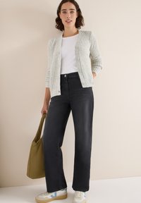 Hellgraue Strickjacke mit strukturiertem Muster, getragen über einem weißen Oberteil. Dunkelblaue weite Jeanshose und weiße Sneakers mit blauen Akzenten.
