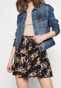 Veste en denim à lavage clair, boutonnée à l'avant, avec deux poches poitrine, accompagnée d'une jupe plissée noire à fleurs, ornée de fleurs roses et jaunes vives.