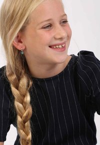 Meisje met gevlochten blond haar, gekleed in een zwart shirt met verticale witte strepen en kleine hangende oorbellen met een turquoise detail.