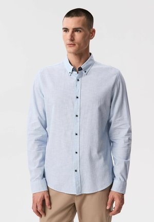 Młody mężczyzna o krótkich włosach, ubrany w jasnoniebieską koszulę zapinaną na czarne guziki oraz beżowe spodnie, stojący na jednolitym tle.