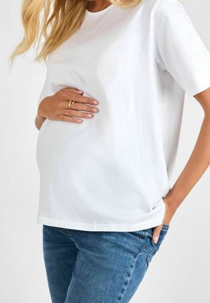 Kobieta w ciąży, ubrana w biały t-shirt i niebieskie dżinsy, delikatnie trzyma brzuch jedną ręką, druga ręka w kieszeni.