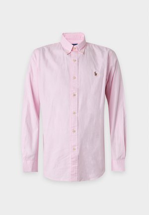 Jeune homme portant une chemise rose boutonnée, un jean bleu clair, des baskets blanches et une casquette de baseball marron, debout devant un fond blanc.