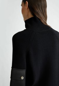 Maglione nero lavorato a maglia con colletto alto, presenta una texture a coste e un polsino in tessuto contrastante impreziosito da un dettaglio con bottone metallico.