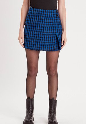 Kort mini-rok in blauw en zwart houndstooth met een zijsplit en knoopdetail, gemaakt van gestructureerde stof. Draag het met zwarte kousen en laarzen.