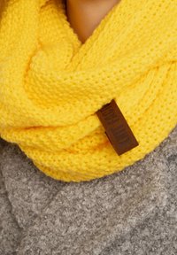 Gele gebreide infinity sjaal met een zachte textuur, voorzien van een bruin lederen label. Draagt over een grijze, gestructureerde kledingstuk.