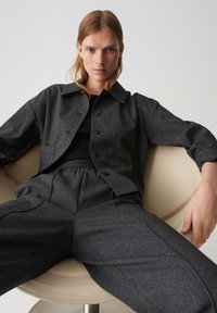 Junge Person mit glattem blonden Haar, die eine dunkelgraue karierte Jacke und Hose trägt, sitzt auf einem beigen modernen Stuhl und schaut nach vorne.