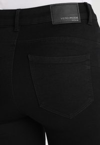 Vaqueros de mezclilla negra con cintura de altura media, un bolsillo trasero de parche y una etiqueta de cuero con marca en la cinturilla. Textura lisa y uniforme.