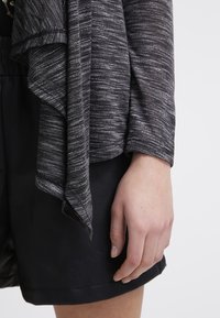 Cardigan in tessuto a maglia grigio e nero con orlo asimmetrico e design morbido e drappeggiato, indossato sopra una gonna in pelle nera liscia.