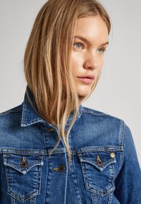 Džinsinė striukė su klasikiniu dizainu, mėlynos spalvos, dviem krūtinės kišenėm su rudos spalvos mygtukais ir matoma geltona siūlė.