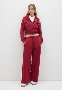 Giacca rossa accorciata con zip e dettagli bianchi abbinata a pantaloni di tuta rossi a gamba larga. Presenta una vita elastica e tasche laterali.