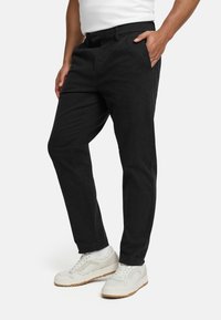 Black cotton trousers with a straight fit, featuring side pockets, a button closure, and worn with white sneakers. Smooth, matte texture.