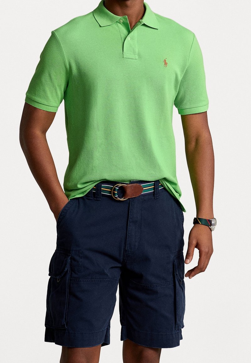 Polo vert en tissu tricoté avec un col et des manches courtes, associé à un short cargo marine avec des poches latérales et une ceinture à motif.