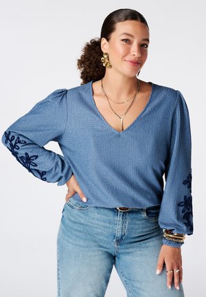 Femme aux cheveux bouclés en queue de cheval portant un chemisier bleu texturé avec une broderie florale sur les manches et un jean bleu clair, souriant légèrement.
