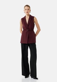 Bordeaux mouwloze blazer met een ceintuur in de taille, scherpe revers en een getailleerde pasvorm, gecombineerd met zwarte wijde broekspijpen en puntige schoenen.
