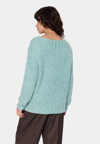 Šviesiai žalias didelis megztinis su reljefiniu tekstūru, plačiu iškirpimu ir nukritusiomis petnešomis, derinamas su laisvomis rudomis kelnėmis.