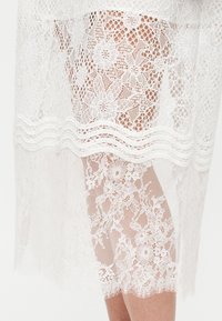 Υφασμα από λευκή δαντέλα με λουλουδένια σχέδια που καλύπτει μερικώς ένα λυγισμένο γόνατο και το κάτω μέρος του ποδιού πάνω σε απλό φόντο.