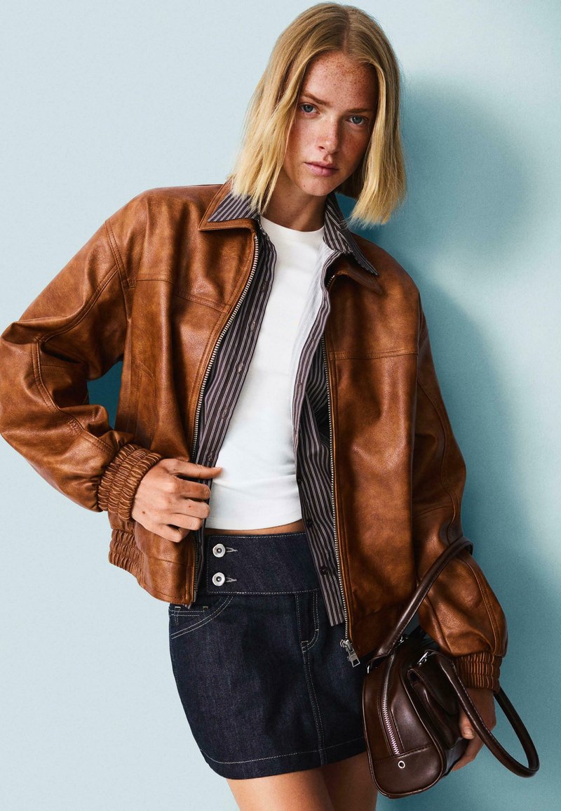 Giubbotto bomber in pelle marrone con polsini a costine e foderato con righe, abbinato a una gonna di jeans con chiusura a doppio bottone.