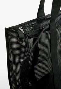 Black mesh tote bag with vertical stripes, thick black handles, top zipper, and a metallic D-ring accent for attaching accessories.
