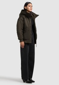 Jachetă puf dintr-o nuanță închisă de verde, cu guler înalt, fermoar frontal și buzunare laterale. Asortată cu pantaloni de denim negri, lejeri și încălțări negre cu toc.